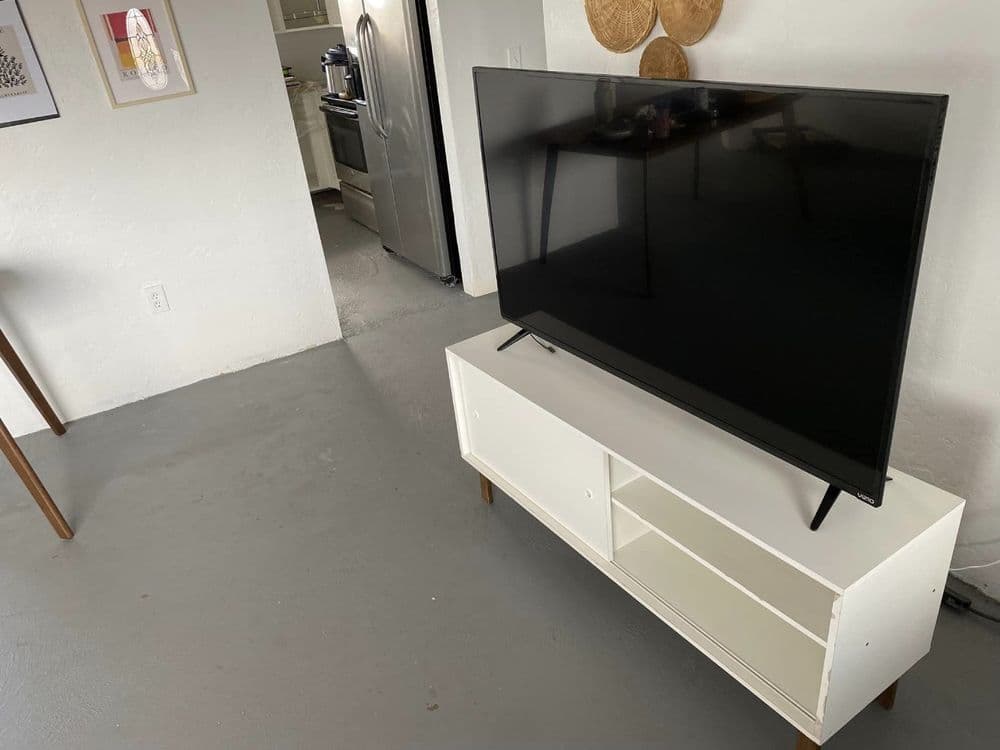 Modern living room with a large TV on a white media console and a glimpse of the kitchen.